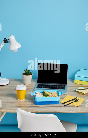 Gesundes Mittagessen mit Reis und Huhn am Arbeitsplatz mit Laptop und Papiere auf Holztisch auf blauem Hintergrund, illustrative Editorial Stockfoto