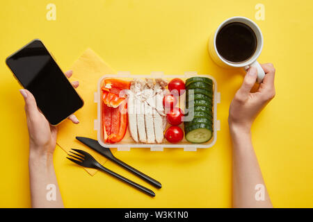 7/8-Ansicht von Frau mit leckeren Mittagessen holding Smartphone und Tasse Kaffee auf gelbem Hintergrund Stockfoto