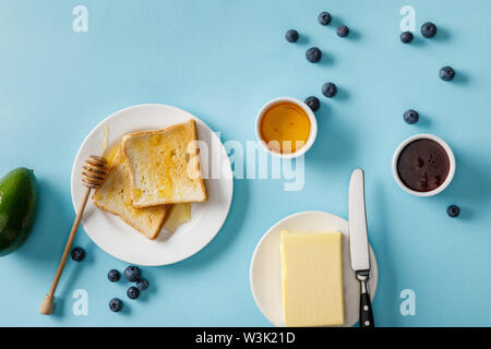 Blick von oben auf die Avocado, Butter, Honig, Marmelade, zerstreut, Blaubeeren und zwei Toasts auf weiße Platten auf blauem Hintergrund Stockfoto