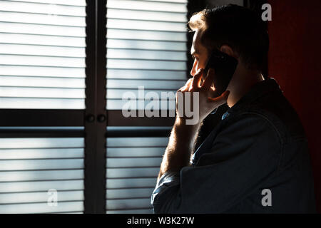 Silhouette der jungen attraktiven Mann Gespräch am Handy beim Stehen in dunklen Raum gegen das Sonnenlicht aus dem Fenster im Schatten Jalousien Stockfoto