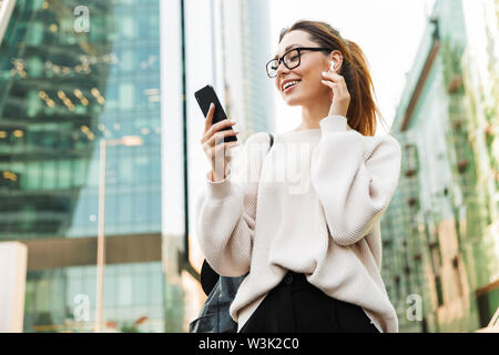 Foto von attraktiven lächelnde Frau tragen Brillen mit Handy und earpod beim Gehen in der grossen Stadt. Stockfoto
