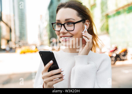 Foto Nahaufnahme von hübsch lächelnde Frau tragen Brillen mit Handy und earpod beim Gehen in der grossen Stadt. Stockfoto