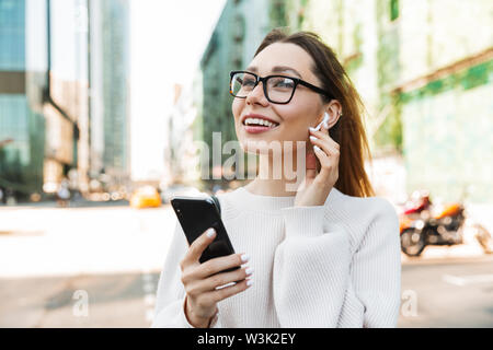 Foto Nahaufnahme von Glücklich lächelnde Frau mit Brille mit Handy und earpod beim Gehen in der grossen Stadt. Stockfoto