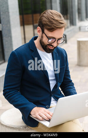 Portrait von optimistisch Geschäftsmann Brillen tragen mit und Suchen am Laptop sitzen im Freien in der Nähe von Gebäude Stockfoto