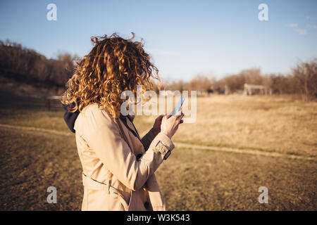 Nicht wiederzuerkennen, junge Frau mit dem lockigen Haar zu tragen beige Mantel stehen draußen in Gelb sonnenbeschienenen Feld an einem warmen Herbsttag mit Ihrem Mobiltelefon. Stockfoto