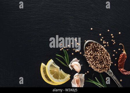 Draufsicht der Koriandersamen in Silber Löffel auf schwarzen Hintergrund mit getrockneten Chili, Zitrone, Kräutern und Knoblauch Stockfoto