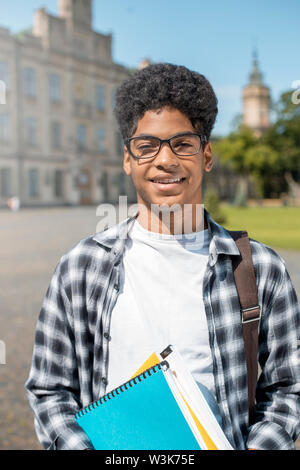 Lächelnder afroamerikanischer Student mit Brille und Büchern in der Nähe des College. Porträt eines fröhlichen schwarzen jungen Mannes, der auf einem Universitätshintergrund steht. Stockfoto