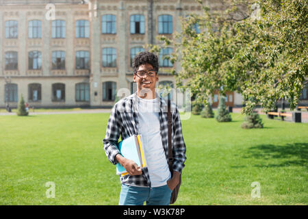 Lächelnder afroamerikanischer Student mit Brille und Büchern in der Nähe des College. Porträt eines fröhlichen schwarzen jungen Mannes, der auf einem Universitätshintergrund steht. Stockfoto