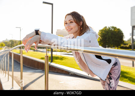 Fröhlicher junger Fitness Mädchen tragen Sportswear Ausgabe Zeit im Freien, sich ausruhen, lehnte sich auf Schienen Stockfoto