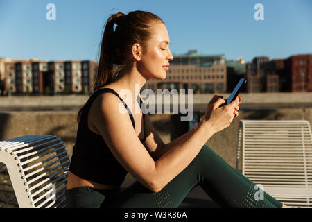 Bild der Sonnenbeschienenen sportliche Frau 20 s tragen Trainingsanzug holding Smartphone und sitzen auf Stuhl, während Sie Workout in der Stadt. Stockfoto