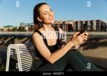 Bild des freudigen sportliche Frau 20 s tragen Trainingsanzug holding Smartphone und sitzen auf Stuhl, während Sie Workout in der Stadt. Stockfoto