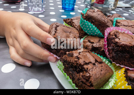 Die Hand eines Kindes nimmt ein Stück Schokoladenkuchen in einem Fall von einer Platte auf eine Tabelle. Stockfoto