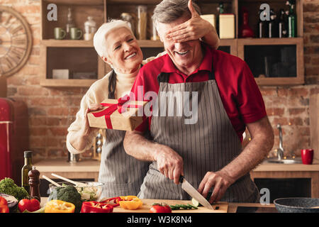 Ältere Frau, Geschenk, das ihr liebevoller Ehemann in Küche begeistert Stockfoto