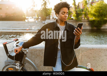 Foto des Denkens afrikanische amerikanische Frau legere Kleidung stehend mit Fahrrad und mit Handy beim Gehen entlang Riverfront Stockfoto