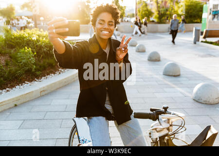 Foto von Glückliche afroamerikanische Frau legere Kleidung lächelnd und unter selfie auf Handy beim Sitzen auf dem Fahrrad im City Park Stockfoto