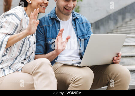Zugeschnittenes Bild der glücklichen jungen erstaunliche paar Geschäftsleute Kollegen draußen im Freien auf Schritte mit Laptop winken im Gespräch mit Fami Stockfoto