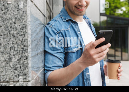 Zugeschnittenes Bild der glücklichen jungen erstaunlichen Mann Geschäftsmann posiert im Freien außerhalb zu Fuß per Mobiltelefon trinken Kaffee plaudern. Stockfoto