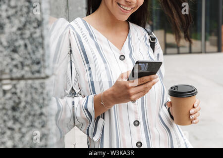 Zugeschnittenes Bild der glücklichen jungen schönen Frau Geschäftsmann posiert im Freien außerhalb zu Fuß per Mobiltelefon trinken Kaffee plaudern. Stockfoto