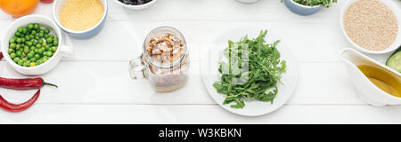 Ansicht von oben Glas Glas mit Muttern in der Nähe von Platte mit grünen Rucola auf hölzernen weissen Tisch, Panorama-Aufnahme ein. Stockfoto