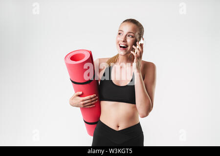 Bild von aufgeregt, sportliche Frau mit Trainingsanzug tragen Fitness Matte und Gespräch am Handy während Aerobic über weiße Wand isoliert Stockfoto