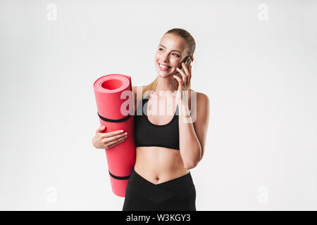 Bild der glücklichen sportliche Frau mit Trainingsanzug tragen Fitness Matte und Gespräch am Handy während Aerobic über weiße Wand isoliert Stockfoto