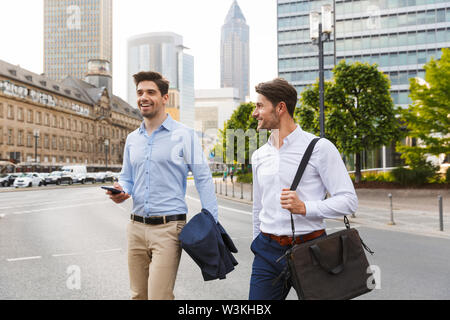 Bild von einem positiven Glücklich lächelnde junge zwei Freunde und Kollegen business men Walking im Freien neben der Suche mit Telefon. Stockfoto
