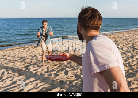 Zwei glückliche junge Männer spielen mit Frisbee am Strand, in der Nähe von hinten von einem Mann Stockfoto