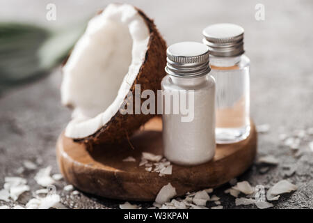 Selektiver Fokus der Flaschen mit Kokosnuss Schönheit Produkte auf Holzbrett in der Nähe von Coconut Hälfte auf Grau strukturierten Hintergrund Stockfoto