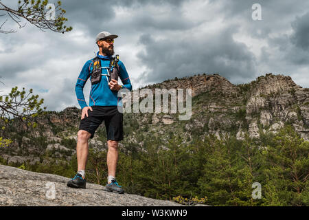 Männliche Läufer stehen auf einem Felsen in den Bergen. Athlet läuft in den Bergen zwischen den Felsen. Mann im blauen Jersey und schwarze Shorts Training im Freien Stockfoto