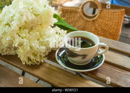 Eine Tasse schwarzen Kaffee auf einem Holztisch und ein Stroh Beutel mit hydragea Blumen auf den Hintergrund. Stockfoto