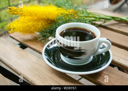 Eine Tasse schwarzen Kaffee auf einem Holztisch und ein Stroh Beutel mit hydragea Blumen auf den Hintergrund. Stockfoto
