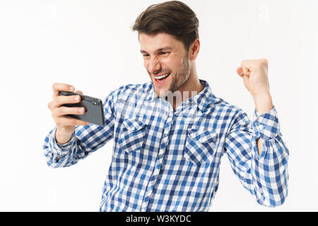 Foto von erfolgreicher Mensch in casual Shirt spielen video game auf Handy und ballte die Faust auf weißem Hintergrund Stockfoto