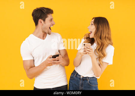 Portrait von attraktiven Paar der Mann und die Frau in der Basic t-shirts sich einander halten Smartphones über Gelb Hintergrund isoliert Stockfoto