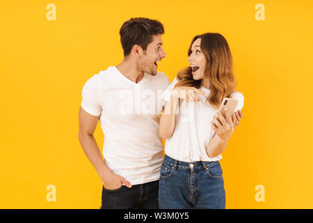 Portrait von schönes Paar der Mann und die Frau in der Basic t-shirts sich einander, während sie Smartphone über Gelb Hintergrund isoliert Stockfoto