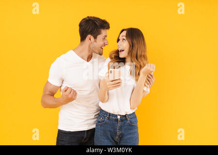 Portrait von freuen Paar der Mann und die Frau in der Basic t-shirts sich einander, während sie Smartphone über Gelb Hintergrund isoliert Stockfoto