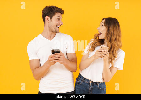 Portrait von schönes Paar der Mann und die Frau in der Basic t-shirts sich einander halten Smartphones über Gelb Hintergrund isoliert Stockfoto