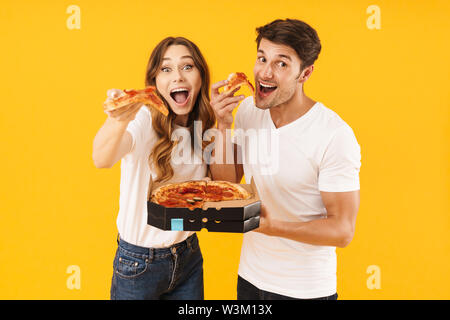 Portrait von freundlichen Paar der Mann und die Frau in der Basic t-shirts Lächeln beim essen Pizza aus, über Gelb Hintergrund isoliert Stockfoto