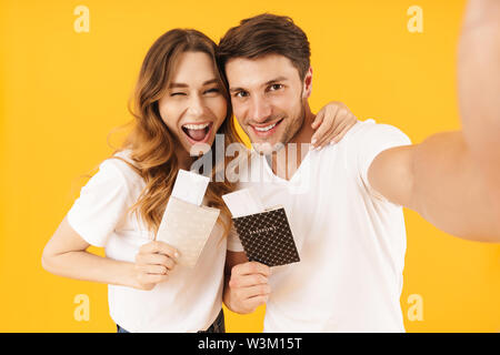 Portrait von lächelnden Paar der Mann und die Frau in der Basic t-shirts Holding Reisen Tickets mit Pässen während der Einnahme von selfie Foto über Gelb backgr isoliert Stockfoto