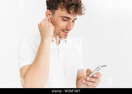 Foto Nahaufnahme des freudigen curly Mann in casual t-shirt lächelnd, während Sie Ohrhörer und Holding Smartphone auf weißem Hintergrund Stockfoto