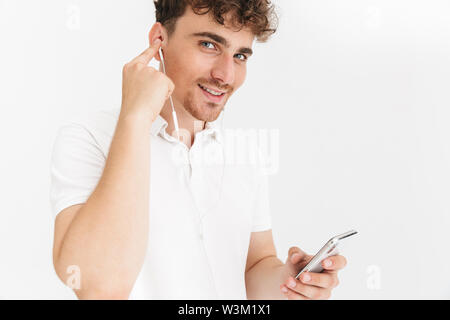 Foto closeup von Happy curly Mann in casual t-shirt lächelnd, während Sie Ohrhörer und Holding Smartphone auf weißem Hintergrund Stockfoto