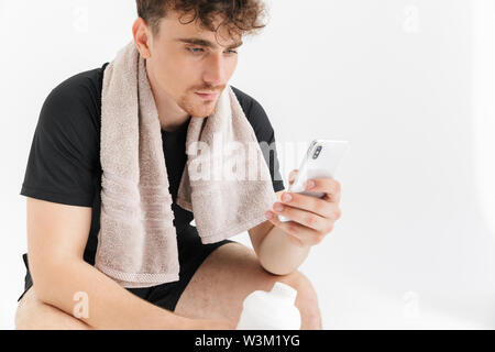Foto Nahaufnahme der Brünette junge Mann mit Handtuch sitzt auf einem Stuhl und Sie Ihr Smartphone nach dem Workout isoliert auf weißem Hintergrund Stockfoto