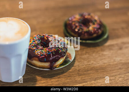 Schlechtes Frühstück. Kaffee mit einem gezeichneten Herz und Milch auf einen hölzernen Tisch in einem Coffee Shop. zwei Schokolade Donuts mit Streuung auf den Tisch neben der Co Stockfoto