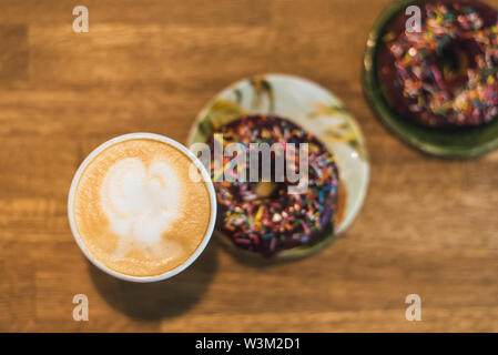 Schlechtes Frühstück. Kaffee mit einem gezeichneten Herz und Milch auf einen hölzernen Tisch in einem Coffee Shop. zwei Schokolade Donuts mit Streuung auf den Tisch neben der Co Stockfoto