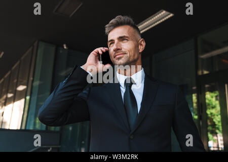 Foto von einem optimistischen ältere Grauhaarige stattlichen business Mann draußen auf der Straße in der Nähe von Business Center im Gespräch per Handy. Stockfoto