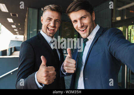 Foto der freudigen zwei Geschäftsleute im Anzug mit Daumen nach oben und unter selfie beim Stehen außerhalb von Job Center gekleidet Stockfoto