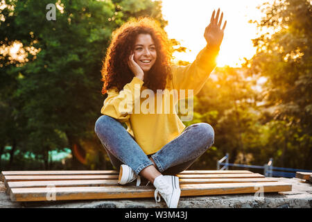 Foto eines glücklichen süße junge Studentin lockiges Mädchen sitzt auf der Bank draußen in der Natur Park mit schönen Sonnenlicht beiseite, winken zu Freunden. Stockfoto