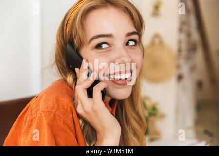 Bild eines nett lächelnden jungen blonden Mädchen zu Hause drinnen sprechen per Handy sitzen auf Stuhl. Stockfoto