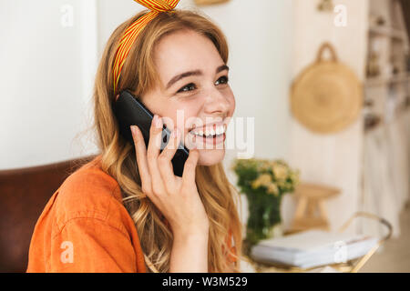 Bild von einem niedlichen emotionale glückliche junge blonde Mädchen zu Hause drinnen sprechen per Handy sitzen auf Stuhl. Stockfoto