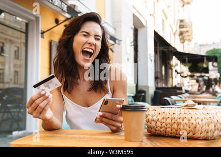 Portrait von Angeregten cute woman mit Stroh Beutel, Kreditkarte und Handy während der Sitzung in gemütliches Cafe im Freien Stockfoto