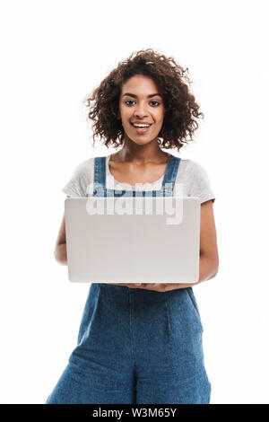 Bild des schönen afrikanischen amerikanischen Frau in Jeans Overalls und lächelnd Laptop auf weißem Hintergrund gekleidet Stockfoto